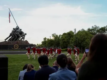 The Marine Corps War Memorial stands under a clear sky as a marching band in red colonial-style jackets performs on the lawn in front of it, with spectators nearby.