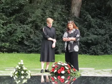 Two women stand beside the Memorial to the Murdered Sinti and Roma of Europe, next to a dark reflective water basin with floral wreaths.