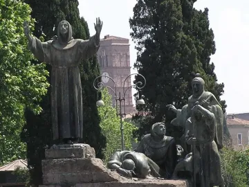 Bronze Monument to Saint Francis of Assisi with robed figures on a stepped stone plinth under a blue sky.