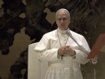 A man in white papal attire stands at a microphone with his hands clasped before a dark textured wall.