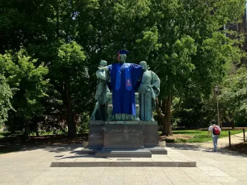 The Alma Mater statue with a central bronze figure in a blue graduation gown, flanked by two figures on a stone pedestal.