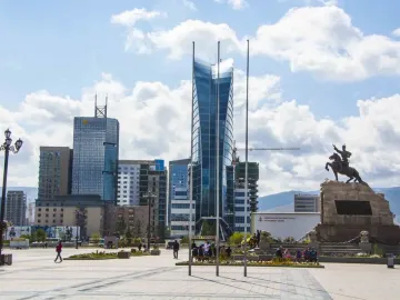 Statue of Damdin Sükhbaatar on a stone plinth in a paved square with tall glass skyscrapers behind it.