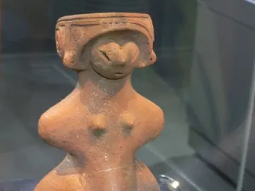 Jōmon Venus clay figurine with a headdress, slit eyes, and rounded body on a dark display base.
