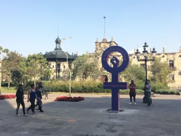 Antimonumenta, a purple pillar-like sculpture with a circular loop and raised fist, stands in a public square with pedestrians and a historic building in the background.