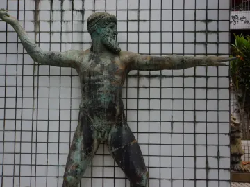 Artemision Bronze sculpture of a nude, bearded male figure with raised arm and extended arm, shown against a white tiled wall.