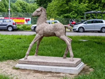 Holbeinpferd foal sculpture stands on a concrete base in grass, with a road, parked vehicles, graffiti-covered fence, and trees behind it.