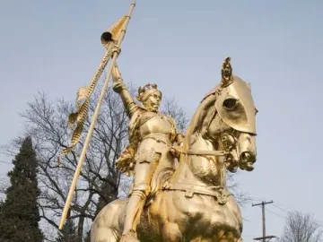 Joan of Arc equestrian statue in golden bronze on a stone pedestal in a park setting.