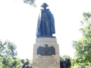 Von Steuben monument with a dark statue atop a stone pedestal, flanked by bronze figures and carved inscriptions.