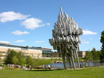 Norra skenet, a large abstract silvery sculpture on a lawn near a pond and modern campus buildings.