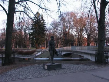 Pushkin statue on a dark pedestal in a park with bare trees and a bridge in the background