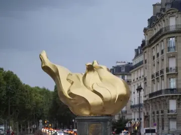 The Flame of Liberty sculpture stands above a memorial of flowers, photos, and locks, with two people nearby and Paris buildings in the background.