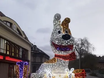 Reksio Monument lit with white and gold lights, with a red scarf and glowing gift boxes nearby.