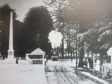 The Needle obelisk stands beside a railway line with a steam train approaching in a tree-lined setting.