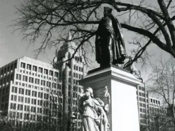 Commodore John Barry statue on a tall pedestal with a relief figure, shown in a park with bare trees and city buildings behind it.