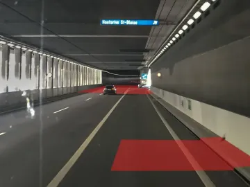 Interior of a brightly lit highway tunnel with a dark car ahead, white lane markings, an overhead Hauterive-St-Blaise sign, and red rectangular overlays on the road.