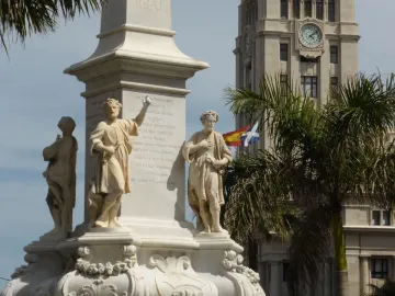 Triunfo de la Candelaria monument with carved figures on a pedestal, with a clock tower, palm trees, and flags in the background.