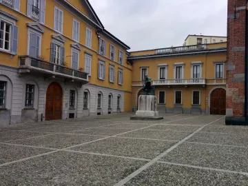Monument to Mosè Bianchi, a dark seated statue on a tall pedestal in a cobblestone piazza.