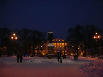 Pushkin statue with an outstretched arm in a snow-covered park at night, with an illuminated building behind it.
