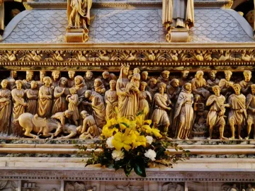 Intricate relief sculpture on the Arca di San Domenico, showing multiple carved figures around a Madonna and Child scene, with flowers in the foreground.