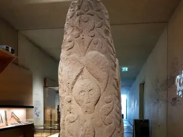 Pfalzfeld Obelisk, a carved sandstone monument with an anthropomorphic face and swirling patterns, displayed on an orange pedestal in a museum corridor.