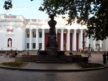 Pushkin bust on a tall pedestal in a paved plaza before a grand columned building.