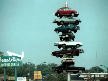 The Spindle sculpture with eight cars stacked vertically on a central pole above a shopping center sign.