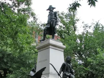 Tadeusz Kościuszko statue on a tall stone pedestal in a park, with bronze figures at the base and trees surrounding it.