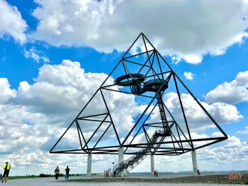 The Tetrahedron steel sculpture stands on tall white pillars with staircases and viewing platforms, with people walking nearby under a blue sky.