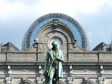 John Cockerill Monument with a standing bronze figure on an ornate pedestal and two seated figures below, in front of a grey stone building.