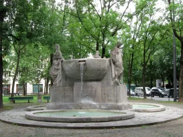 Nornenbrunnen stone fountain with three sculpted figures around a central basin and water flowing into a lower pool.