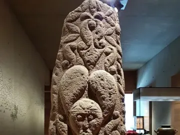 Pfalzfeld Obelisk with a carved human face and swirling motifs in a museum display