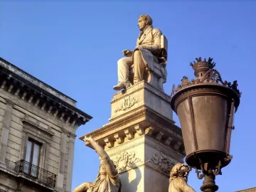 Monument to Vincenzo Bellini with a seated white marble statue on a decorated pedestal, lit by warm sunlight beside a streetlamp.