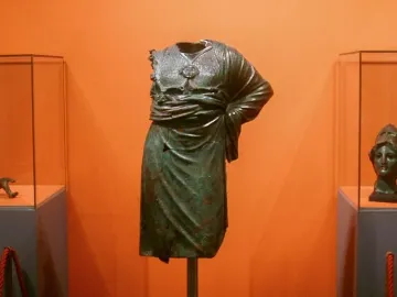 Headless, armless Minerva d\'Arezzo torso on a pole, displayed between museum cases with a detached arm fragment and a helmeted head sculpture.