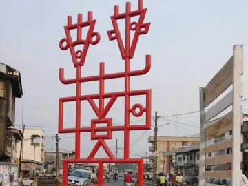 Njé Mo Yé, a bright red abstract sculpture with geometric lines and two stylized figures, stands on a concrete base beside a busy street.