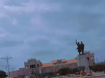 The Demba et Dupont statue stands on a light-colored pedestal near a plaza, with a large building and cloudy sky in the background.