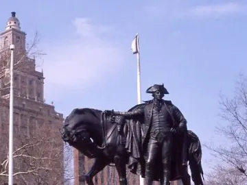 George Washington bronze sculpture beside a harnessed horse on a stone pedestal, with flowers in front and a towered building behind.
