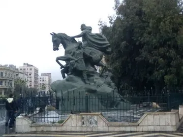 Statue of Saladin, a dark equestrian monument with a cloaked rider on a rearing horse and figures at the base, in an urban square.