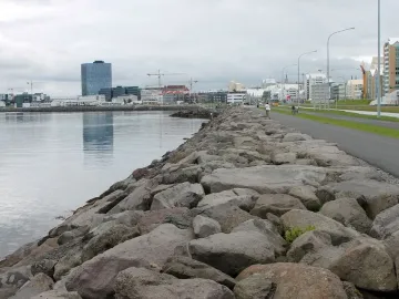 Rocky embankment beside calm water and a paved path with Reykjavik buildings in the distance.