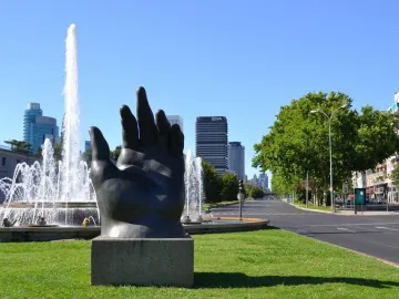 Dark hand sculpture with fingers pointing upward on a stone pedestal beside a fountain and lawn.