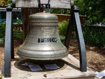 The Liberty Bell with a visible crack and the word “FREEDOM” taped over part of it, resting on a wooden platform with papers below.