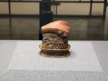 Meat-shaped Stone displayed on an ornate golden stand, resembling a piece of cooked pork belly with a reddish-brown top and mottled grey-brown body.