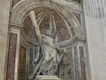 Saint Andrew sculpture standing in an ornate alcove inside St. Peter\'s Basilica, with a carved pedestal and visitors below.