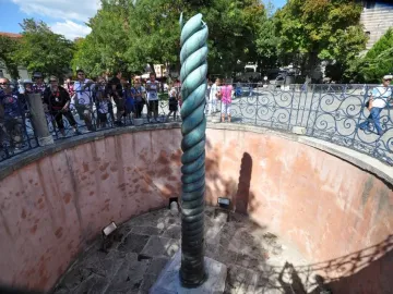 The Serpent Column stands in a sunken pit behind a decorative fence, with visitors gathered around it.