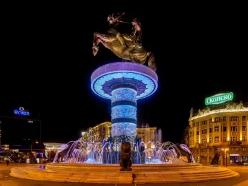 The Warrior on a Horse statue lit in blue and purple above a brightly illuminated fountain with lion sculptures at night.
