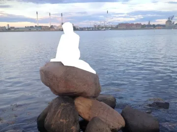 White silhouette of the Little Mermaid seated on rocks by calm water with a distant city skyline
