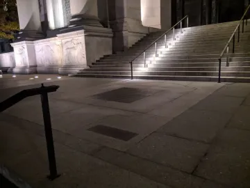 Nighttime view of the American Museum of Natural History plaza with illuminated stone reliefs and inscribed plaques in the foreground.