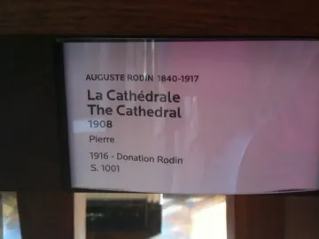 Museum information plaque for Auguste Rodin’s La Cathédrale, with a blurry pink reflection at the right edge.