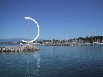 Éole, a large white D-shaped metal sculpture on a rocky breakwater with a marina in the background.