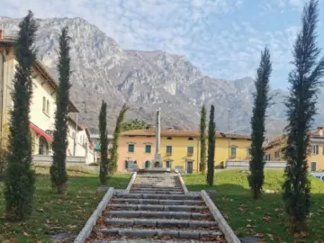 Stone stairs covered with autumn leaves lead up to the Lighthouse of Lierna on a grassy slope, framed by cypress trees and mountains beyond.