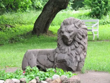 Weathered stone lion sculpture resting on the ground in Mon Repos park, with a tree trunk, bench, and flower bed nearby.